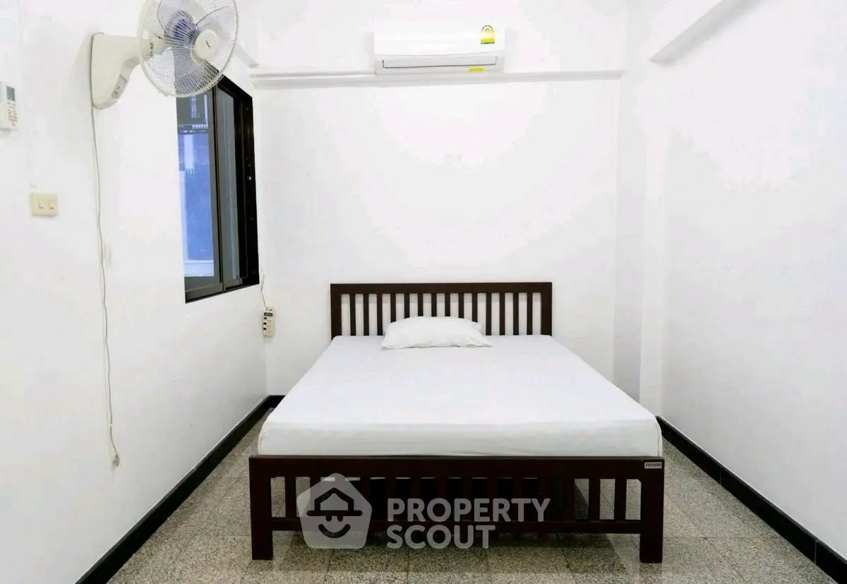 Minimalist bedroom with bed, fan, and air conditioning in a bright, white-painted room.