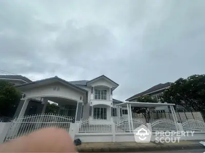 Charming two-story house with white picket fence and lush greenery under a cloudy sky.