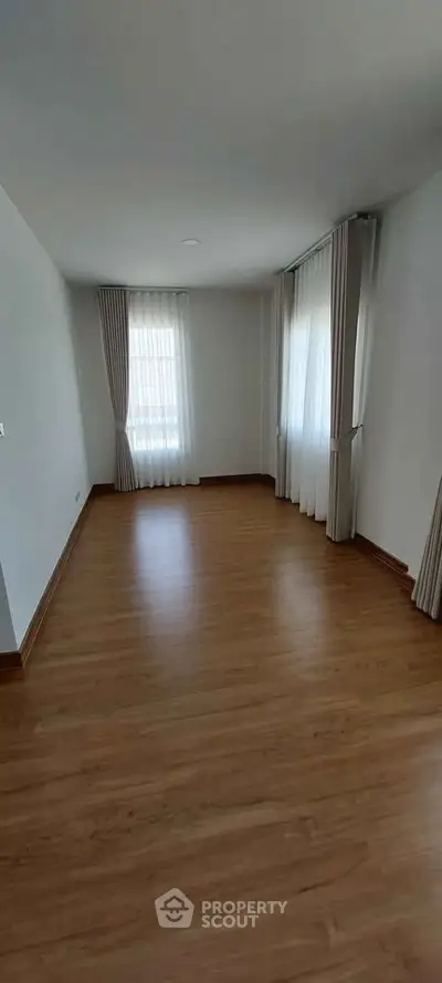 Spacious empty room with wooden flooring and large windows with curtains.