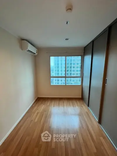 Spacious bedroom with wooden flooring and large window offering city view.
