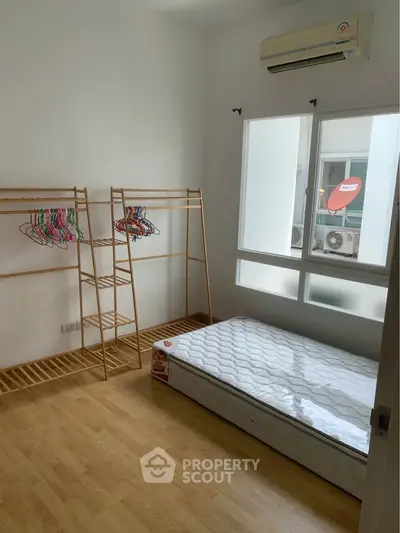 Minimalist bedroom with wooden floor, mattress, and clothes rack, featuring large window and air conditioning.