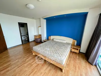 Spacious bedroom with wooden flooring and blue accent wall, featuring a large bed and natural light.