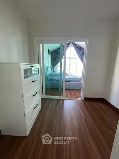 Modern bedroom with wooden flooring and large window, featuring a sleek white dresser and cozy bed.