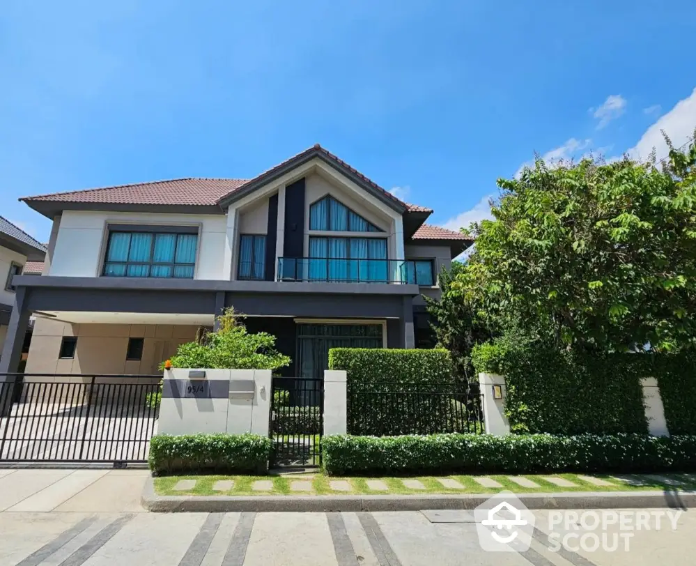 Stunning modern two-story house with lush garden and gated entrance under clear blue sky.