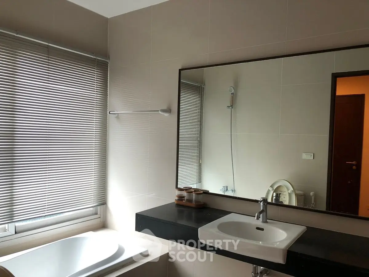 Modern bathroom with sleek sink and bathtub, featuring large mirror and window blinds.