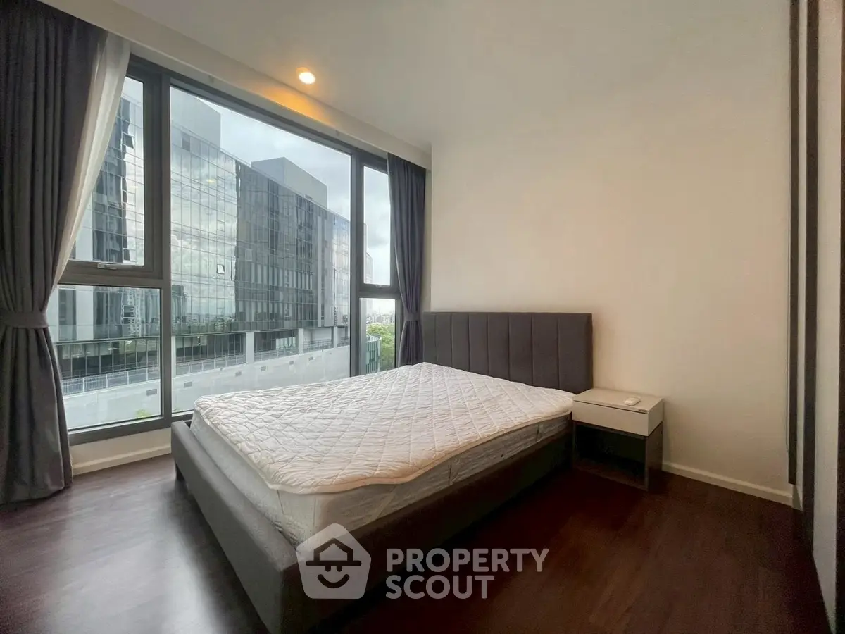 Modern bedroom with large windows and city view, featuring a stylish bed and dark wood flooring.