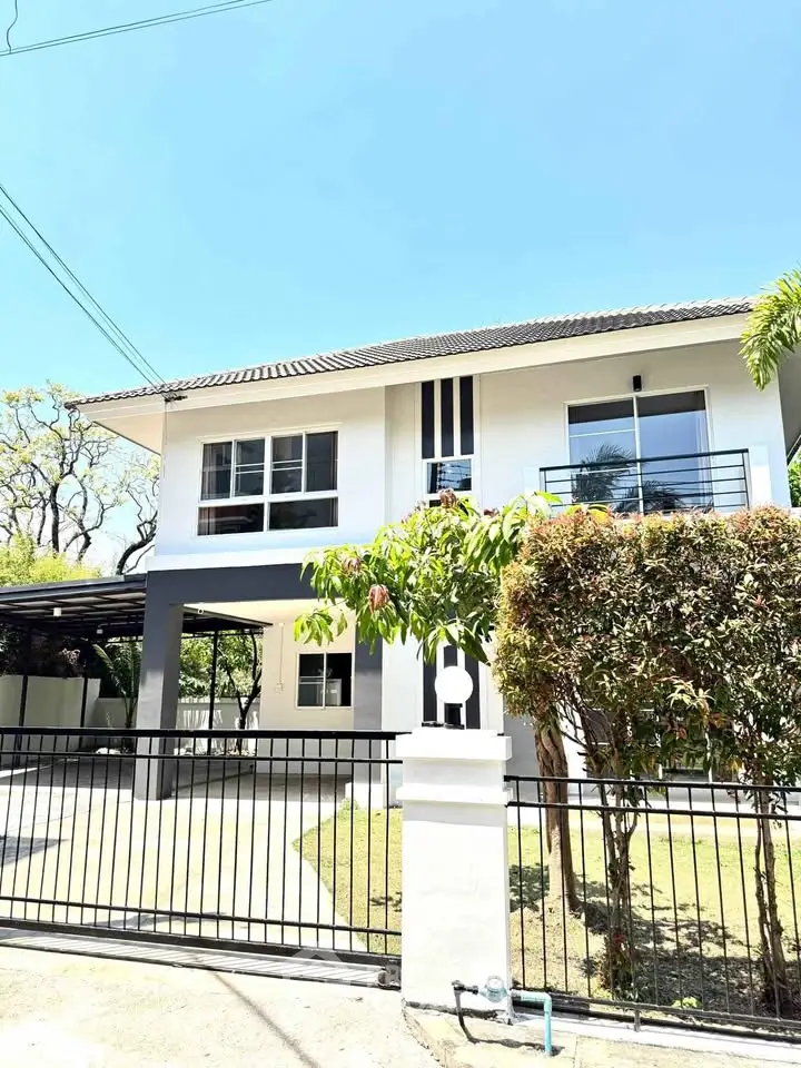 Modern two-story house with spacious balcony and gated entrance, surrounded by lush greenery.