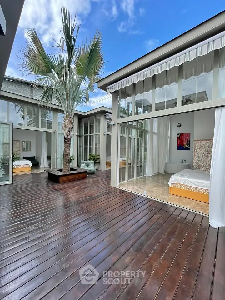 Luxurious modern villa with spacious wooden deck and palm tree, featuring large glass windows and elegant interior design.