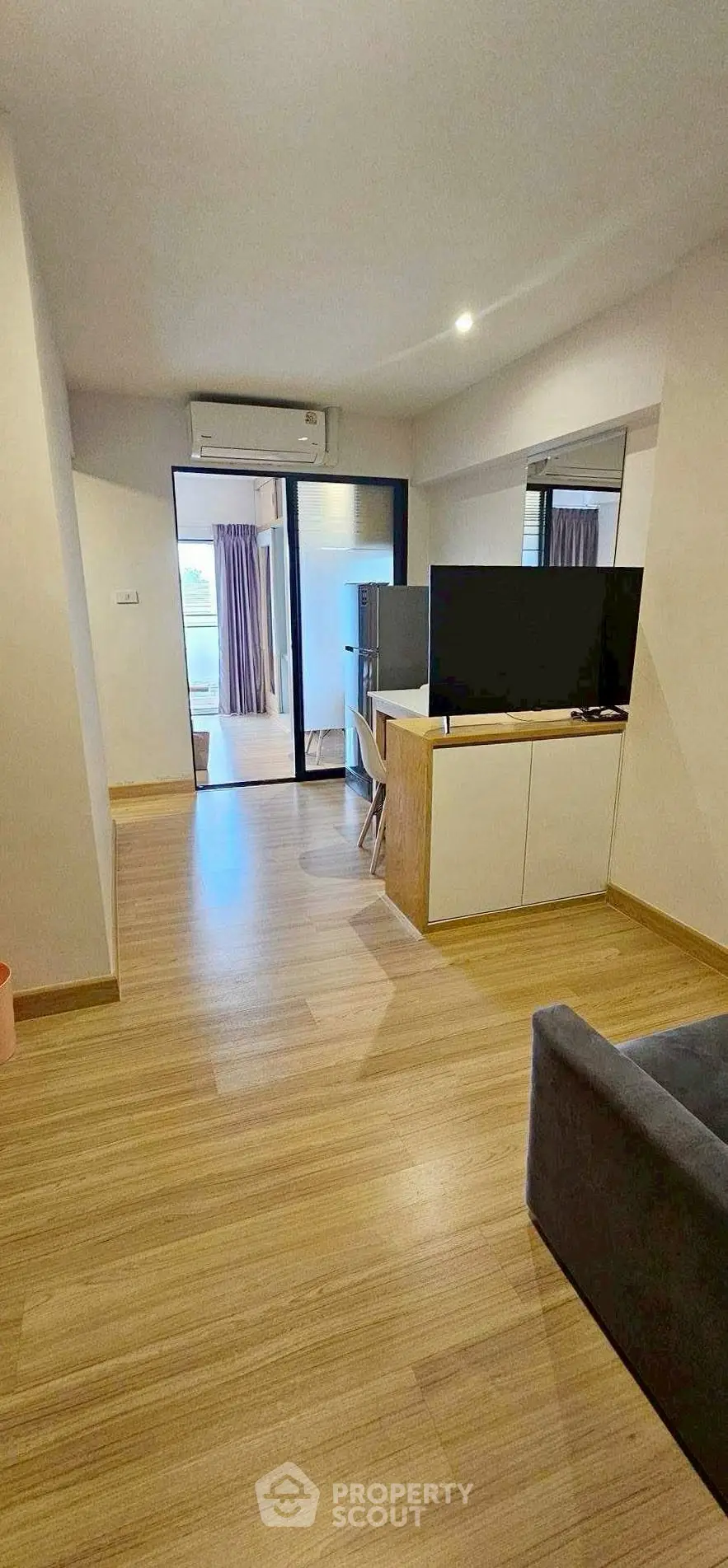 Spacious modern living room with wooden flooring and large TV unit, leading to a balcony with natural light.