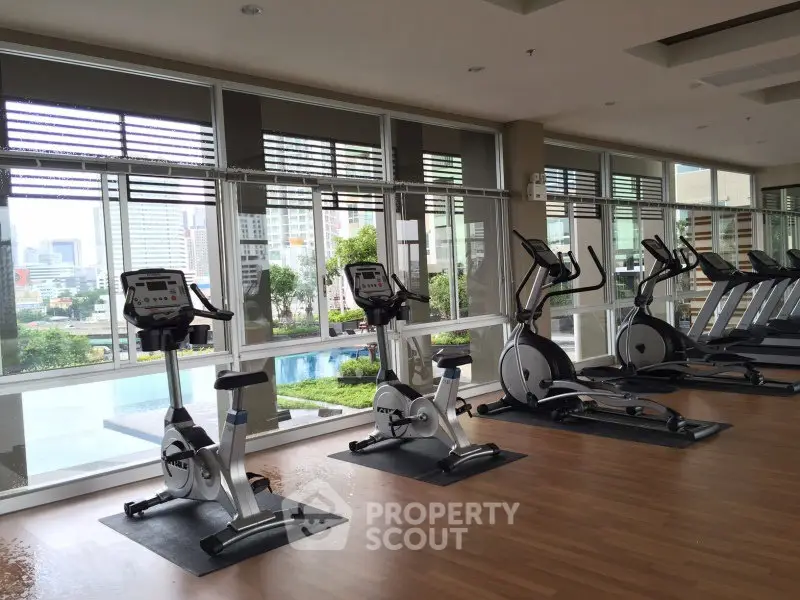 Modern gym with exercise bikes and treadmills overlooking cityscape and pool view.