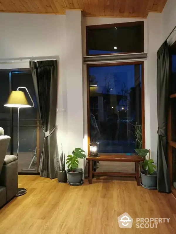 Cozy living room corner with warm wooden floors, large windows offering a night view, and tasteful indoor plants enhancing the serene ambiance.