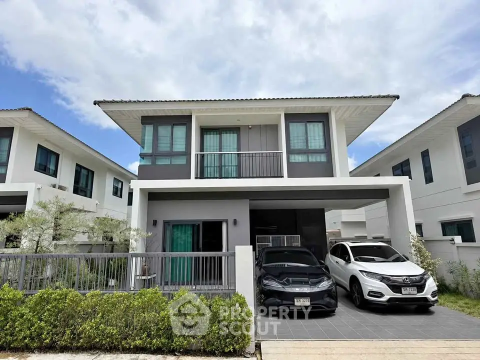 Modern two-story house with driveway and two cars parked, featuring sleek design and lush greenery.