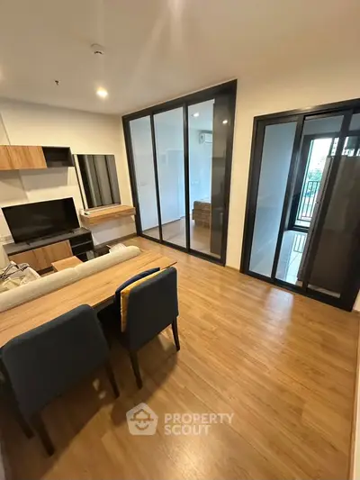 Modern living room with wooden flooring and glass partition, featuring stylish furniture and ample natural light.