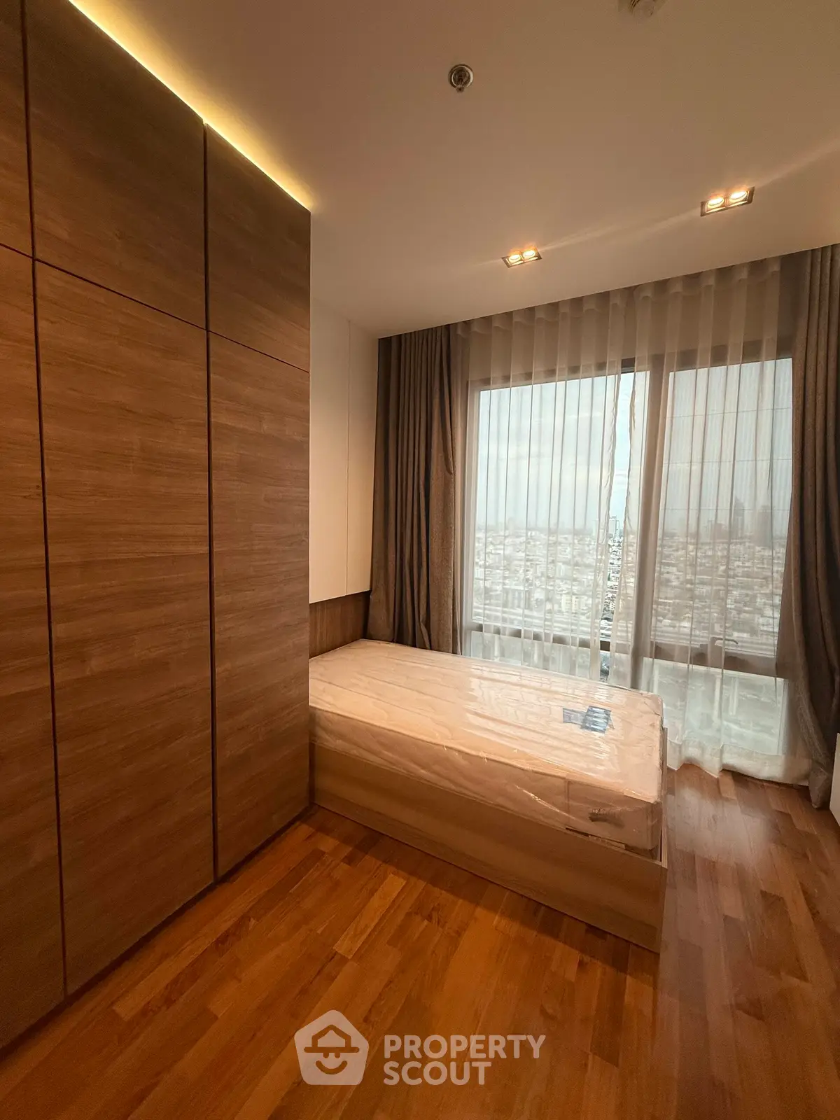 Spacious bedroom with wooden flooring and large window offering city views.