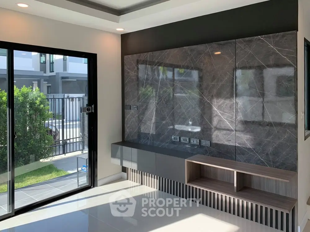 Modern living room with large windows offering ample natural light and a view of the green outdoors, featuring sleek built-in shelving and elegant dark marble wall.