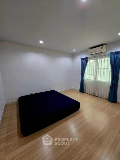 Spacious bedroom with wooden flooring and large window, featuring blue curtains and air conditioning.