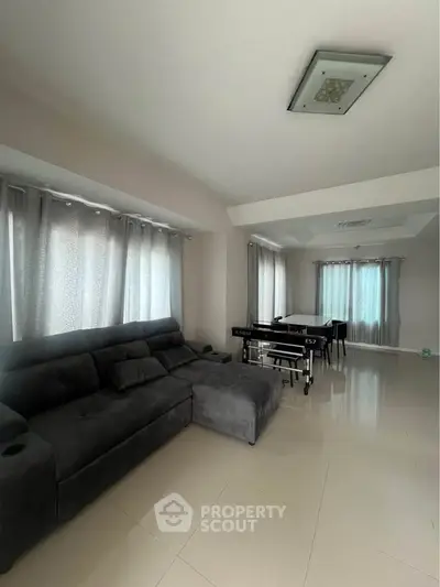 Spacious living room with modern gray sofa and dining area, featuring large windows and elegant curtains.