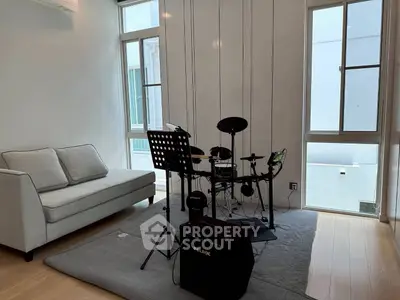 Modern living room with electronic drum set and cozy sofa, featuring large windows for natural light.