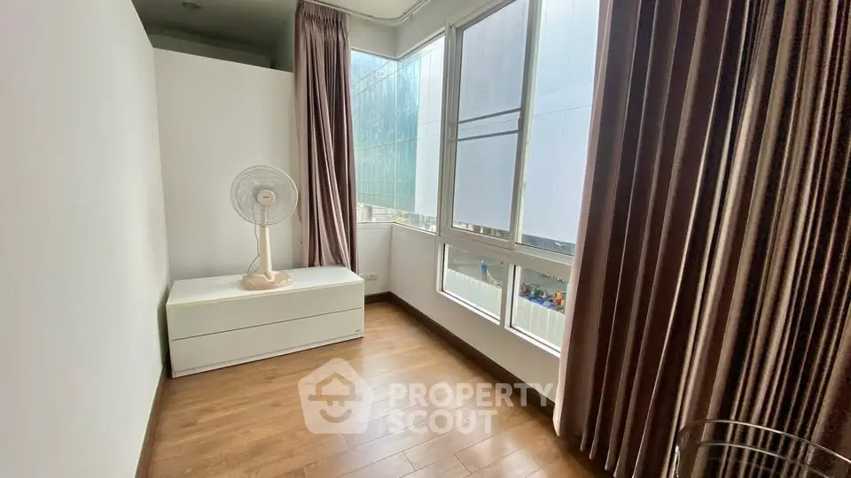 Bright room with large windows and wooden flooring, featuring a fan and curtains.