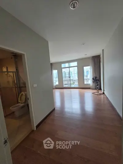 Spacious unfurnished living room with large windows and wooden flooring, adjacent to a bathroom.