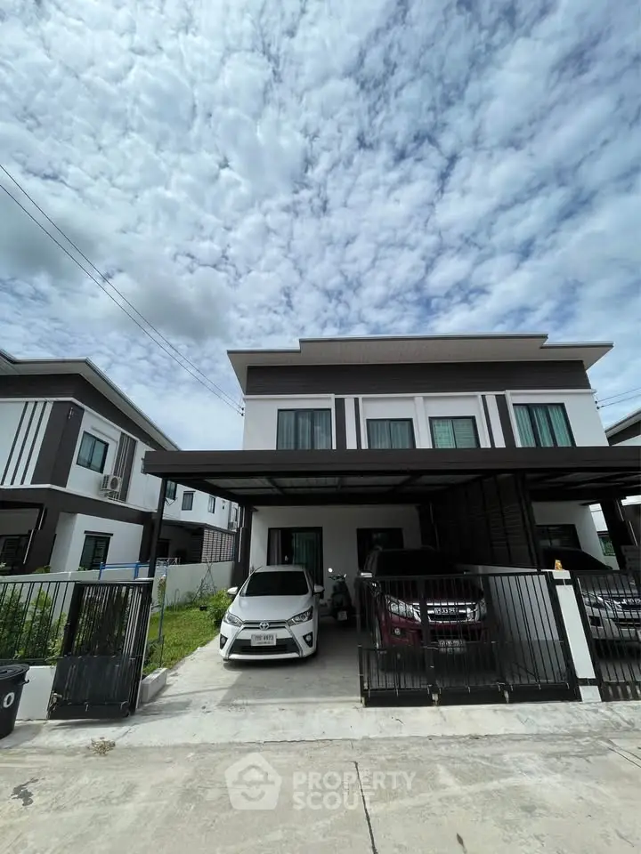 Modern two-story house with driveway and parked cars under a cloudy sky, showcasing contemporary architecture.