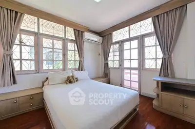 Spacious bedroom with large windows and elegant curtains, featuring a cozy bed and wooden furniture.