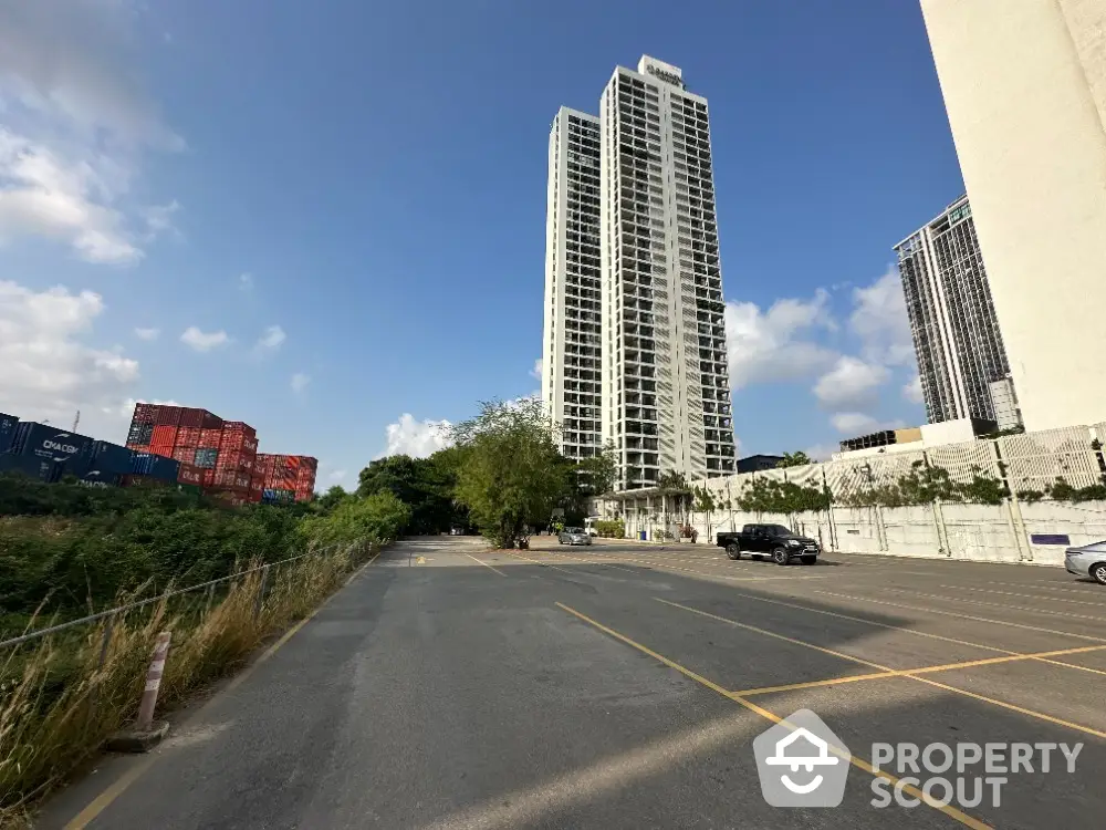 Majestic high-rise condominium towers soar into the blue sky, flanked by lush greenery and a spacious parking area, offering a blend of urban living and nature.