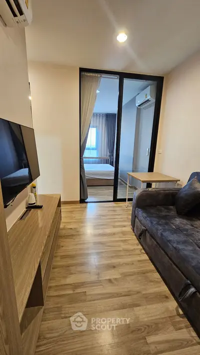 Modern living room with wooden flooring and cozy sofa, leading to a stylish bedroom through glass doors.