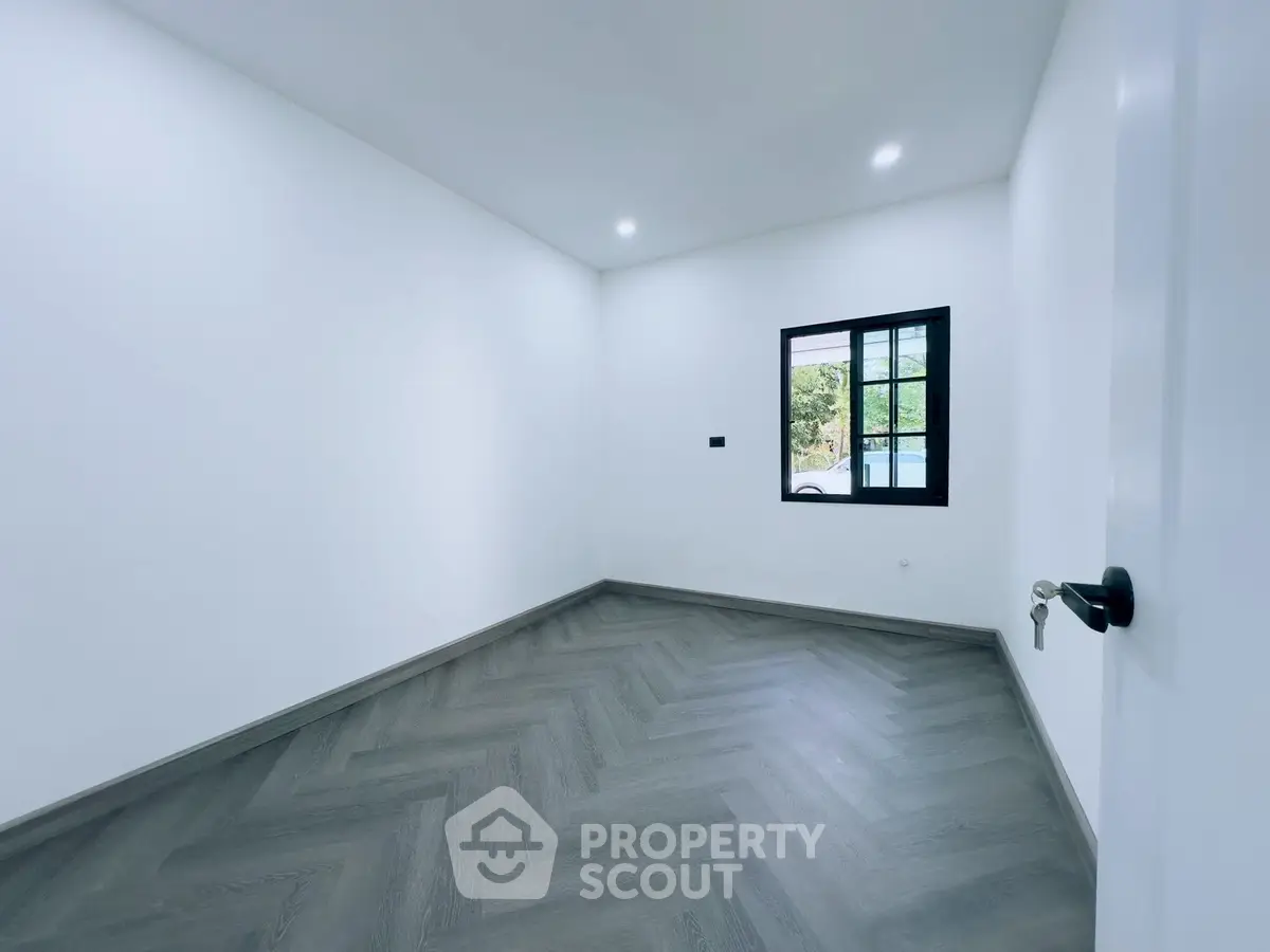 Spacious empty room with elegant herringbone flooring and a large window offering natural light.