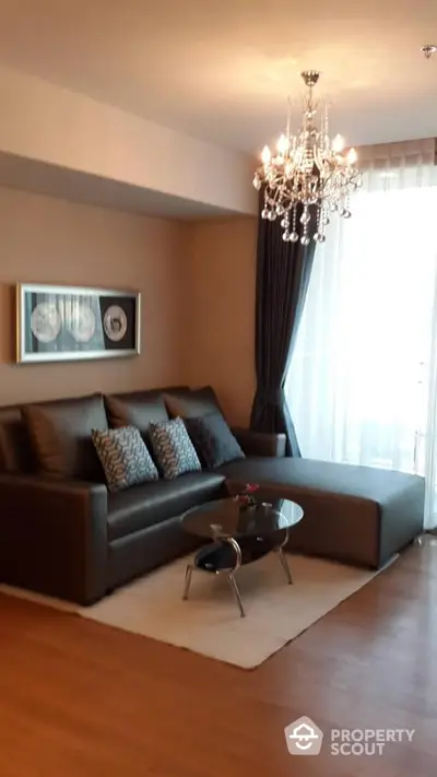 Elegant living room with luxurious chandelier, plush sectional sofa, and modern glass coffee table, set on warm wooden flooring.