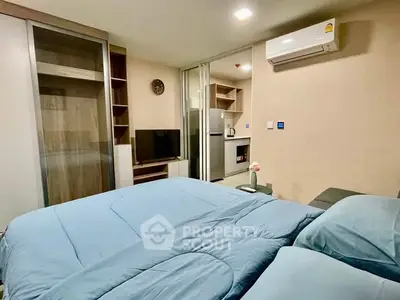 Modern bedroom with blue bedding, TV, and view into a compact kitchen area.