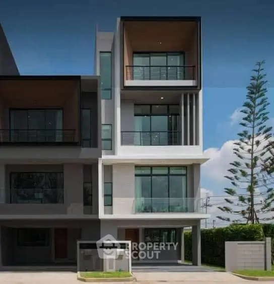 Modern multi-story building with large windows and balconies, surrounded by greenery and clear blue sky.