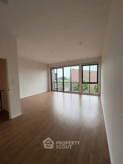 Spacious living room with large windows and wooden flooring, offering natural light and a serene view.