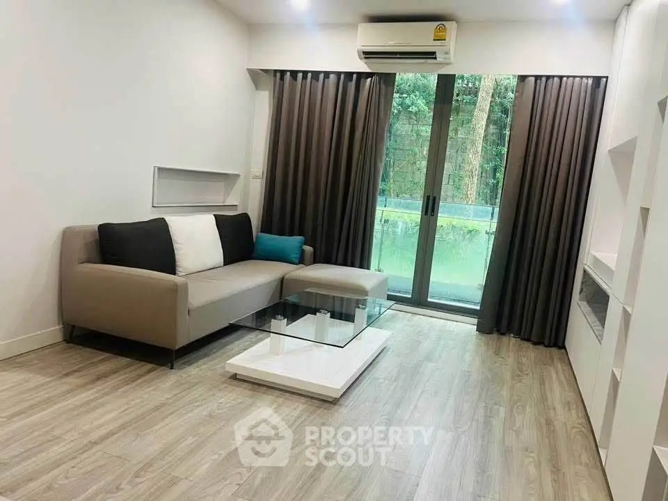 Modern living room with sleek sofa and glass coffee table, featuring large windows and lush greenery view.