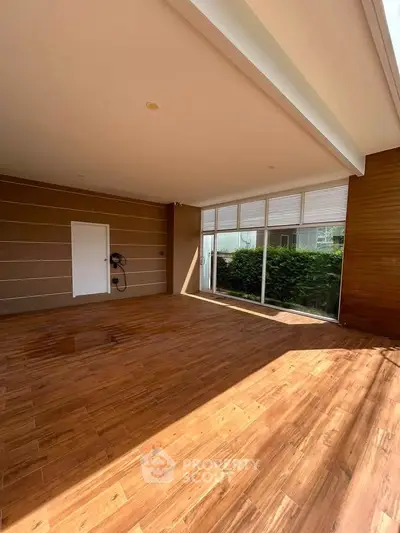 Spacious sunlit room with wooden flooring and large windows overlooking greenery, perfect for modern living.