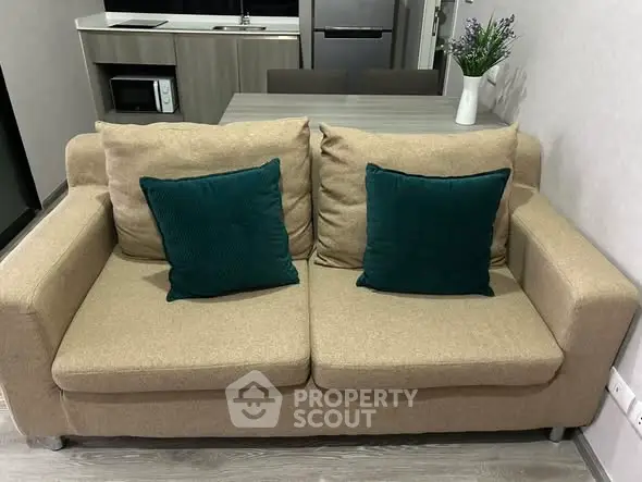 Cozy living room with beige sofa and teal cushions, modern kitchenette in background.