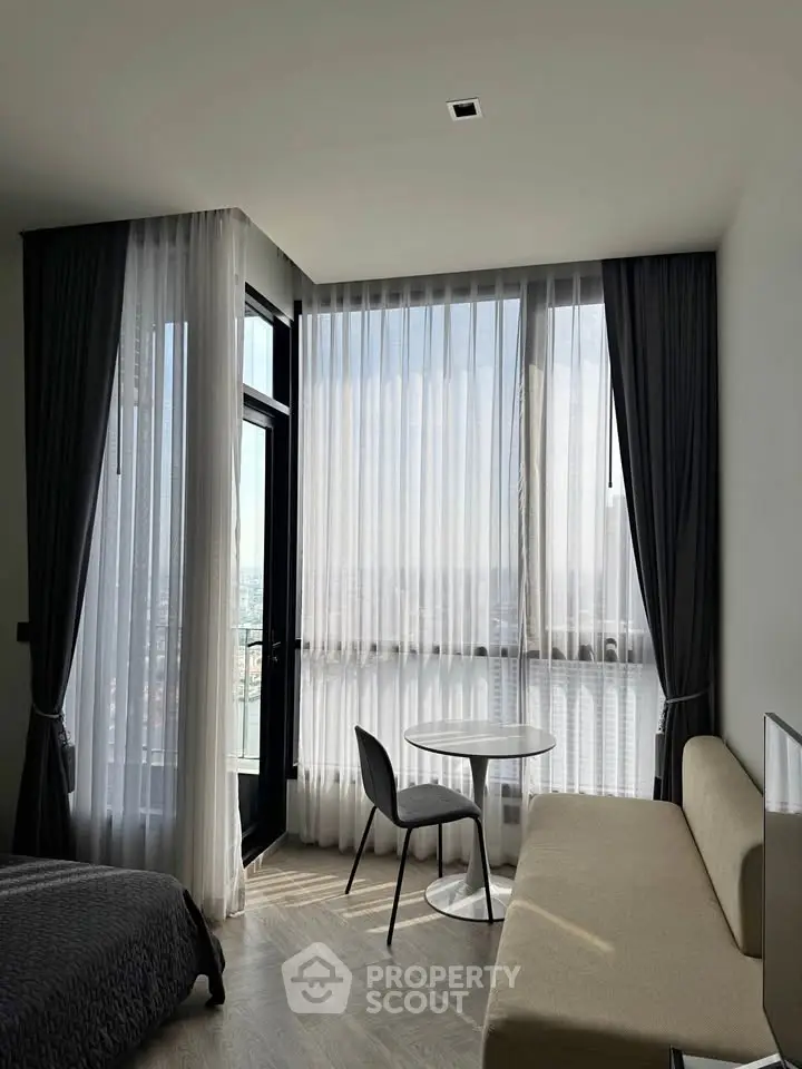 Modern bedroom with floor-to-ceiling windows and elegant curtains, featuring a cozy seating area and a stunning city view.