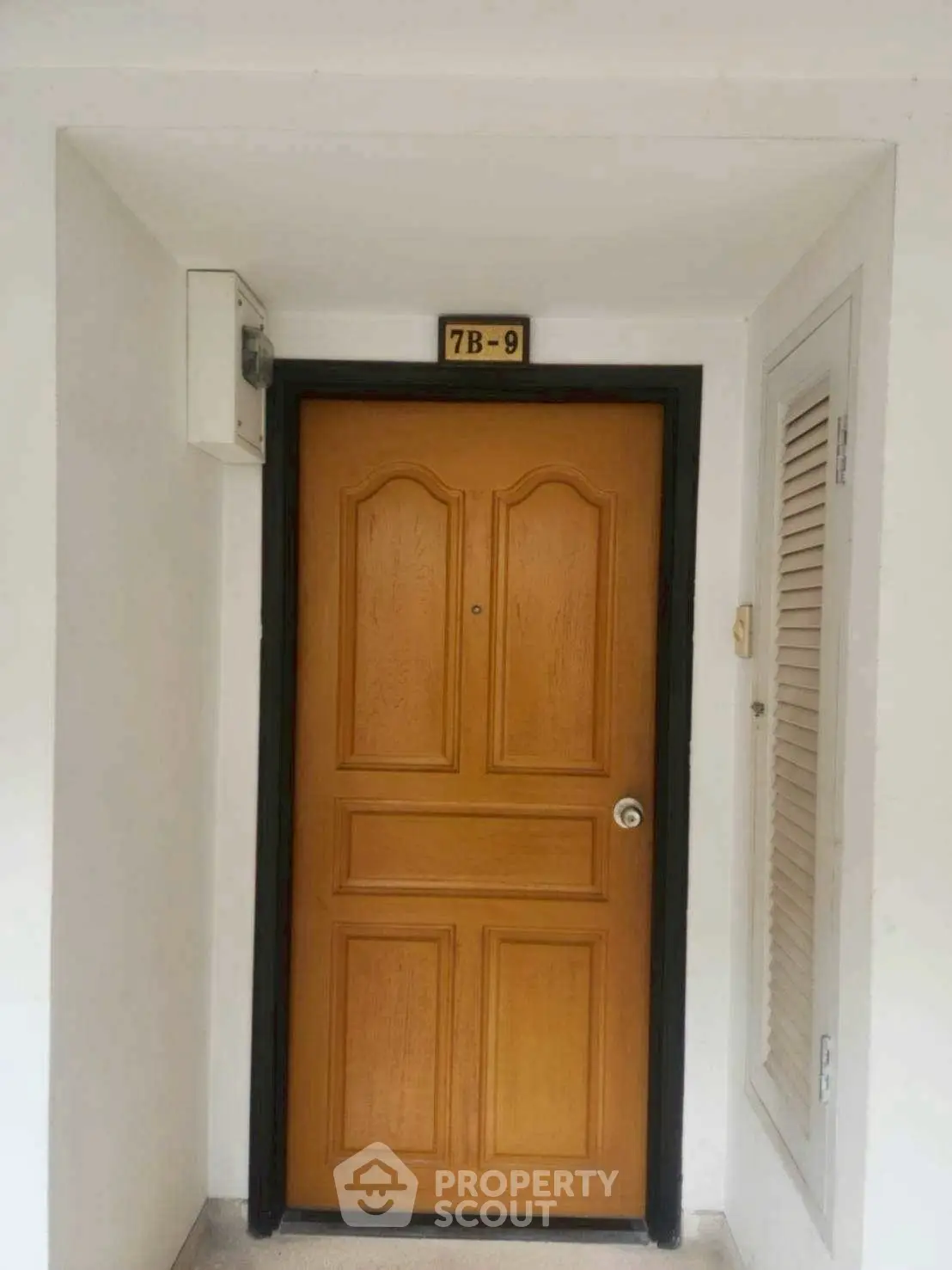 Apartment entrance with wooden door and unit number 7B-9, featuring a simple and clean design.