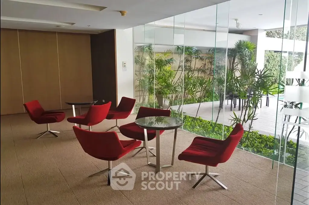 Modern lobby with red chairs and glass walls overlooking a lush garden, perfect for relaxation and meetings.