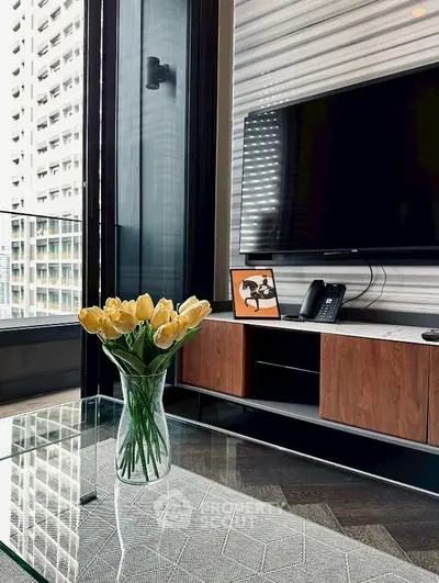 Modern living room with glass table, TV, and city view, featuring elegant decor and natural light.