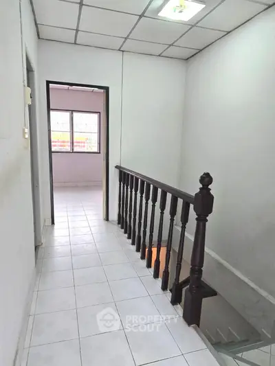 Bright hallway with tiled flooring and wooden railing leading to a room with large windows.