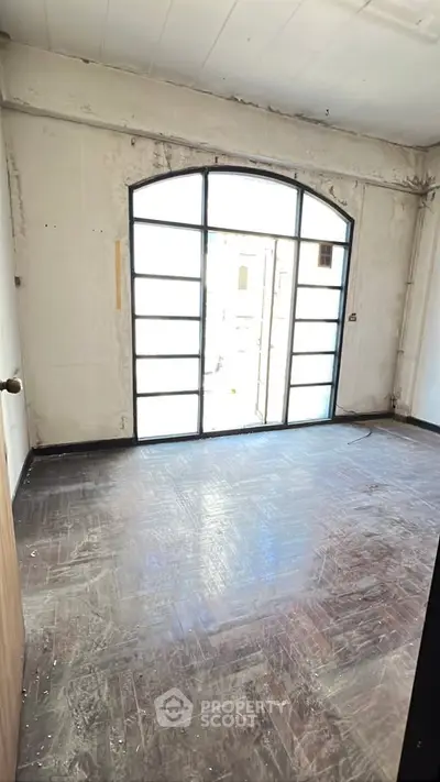 Spacious room with large arched window and wooden flooring, ideal for renovation.