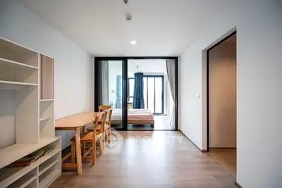 Spacious and modern apartment interior with open floor plan featuring a sleek wooden dining set, ample natural light, and a cozy bedroom visible through the sliding glass door.