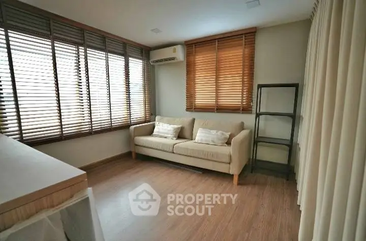 Cozy living room with wooden flooring and large windows with blinds, featuring a comfortable sofa and air conditioning.