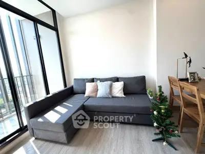 Modern living room with stylish gray sofa and small Christmas tree, featuring floor-to-ceiling windows and wooden dining set.