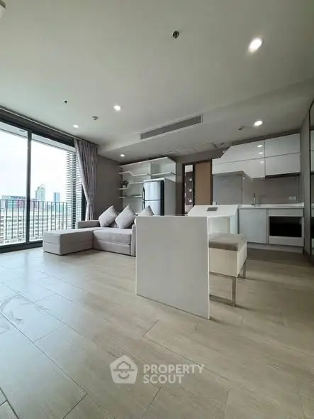 Modern open-plan living room with kitchen, featuring sleek furniture and city view from large windows.