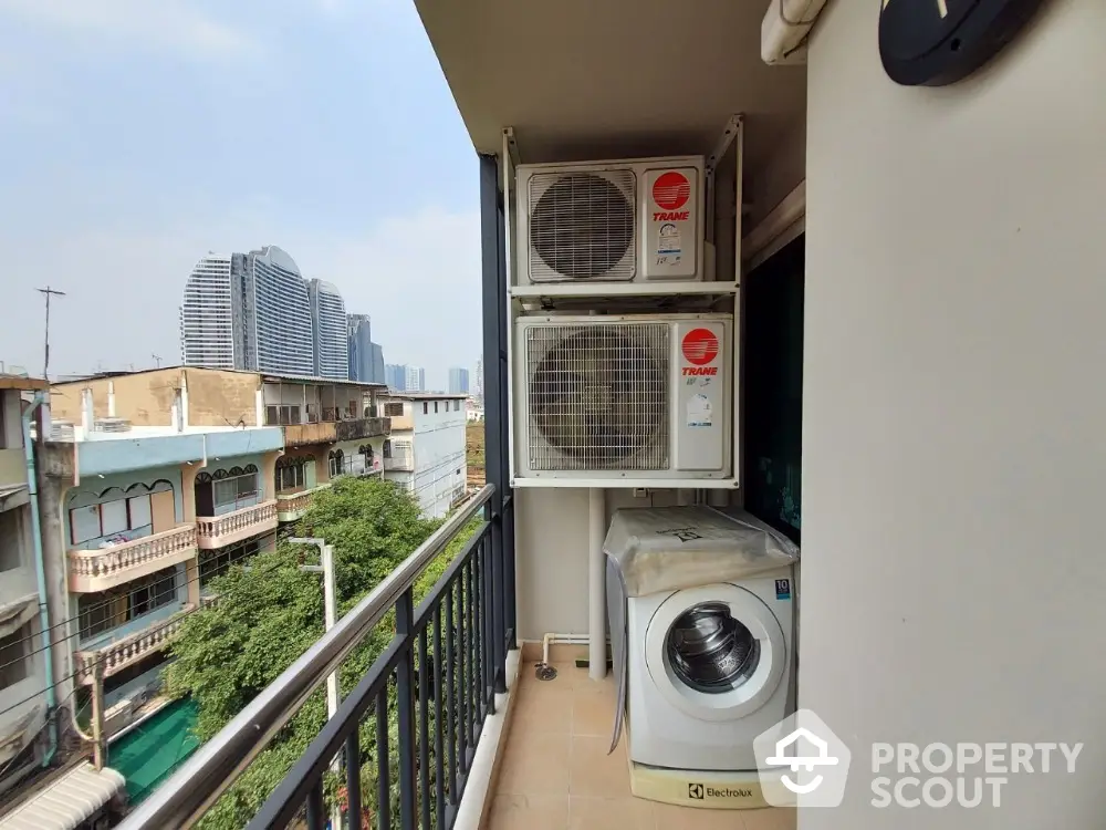 Urban balcony with washing machine and air conditioning units, city view in background.