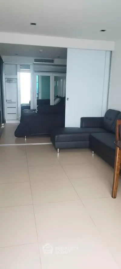 Modern open-plan living room with sleek black sofa and tiled flooring, leading to a stylish bedroom.