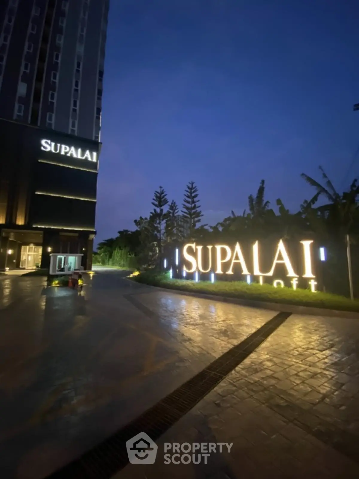 Modern high-rise building entrance at dusk with illuminated signage and lush greenery.