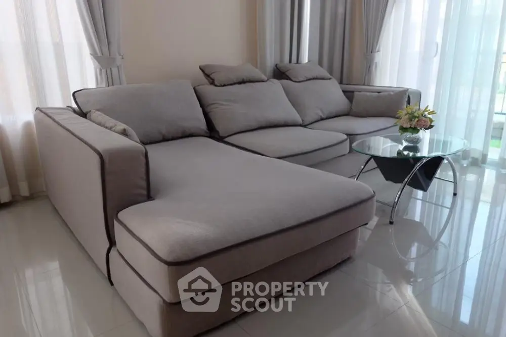 Elegant living room with plush grey sectional sofa, sleek glass coffee table, and airy sheer curtains, radiating modern luxury and comfort.